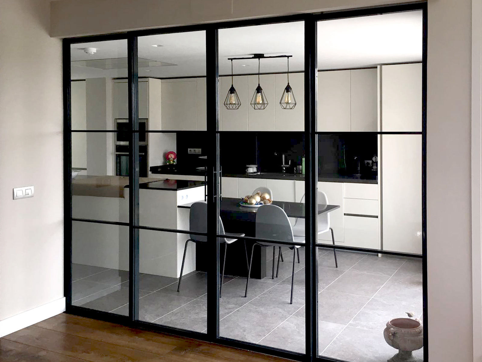 Vista de una cocina moderna con cerramientos de metal y cristal de estilo industrial, que separan el espacio del comedor. La cocina cuenta con muebles blancos de líneas minimalistas, una encimera negra y lámparas colgantes decorativas de diseño geométrico. En el centro, se observa una mesa de comedor con sillas modernas, creando un ambiente funcional y elegante. El suelo es de azulejos grises, complementando la estética contemporánea del espacio.