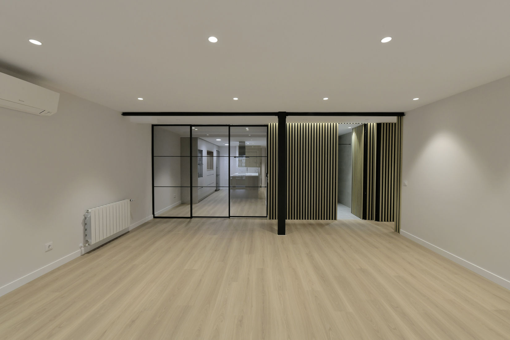 Espacio interior amplio y moderno con suelo de madera clara, delimitado por cerramientos de metal y cristal de estilo industrial que separan la sala principal de la cocina. En el lado derecho, destacan paneles decorativos verticales en negro y dorado, que aportan un toque sofisticado al diseño. Las paredes son de tonos neutros y la iluminación empotrada en el techo crea un ambiente luminoso y acogedor.