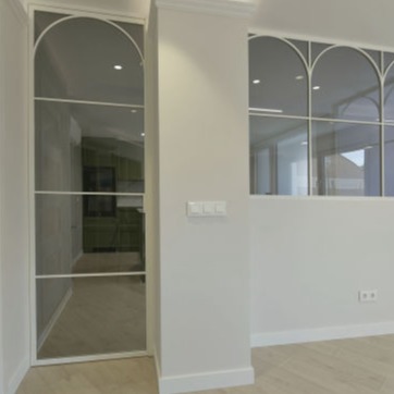 Puerta corredera de diseño moderno con marco metálico blanco y cristal, separando un espacio interior minimalista. A los lados, ventanas acristaladas con arcos decorativos integrados en la estructura, sobre un suelo de madera clara en un ambiente luminoso y elegante.