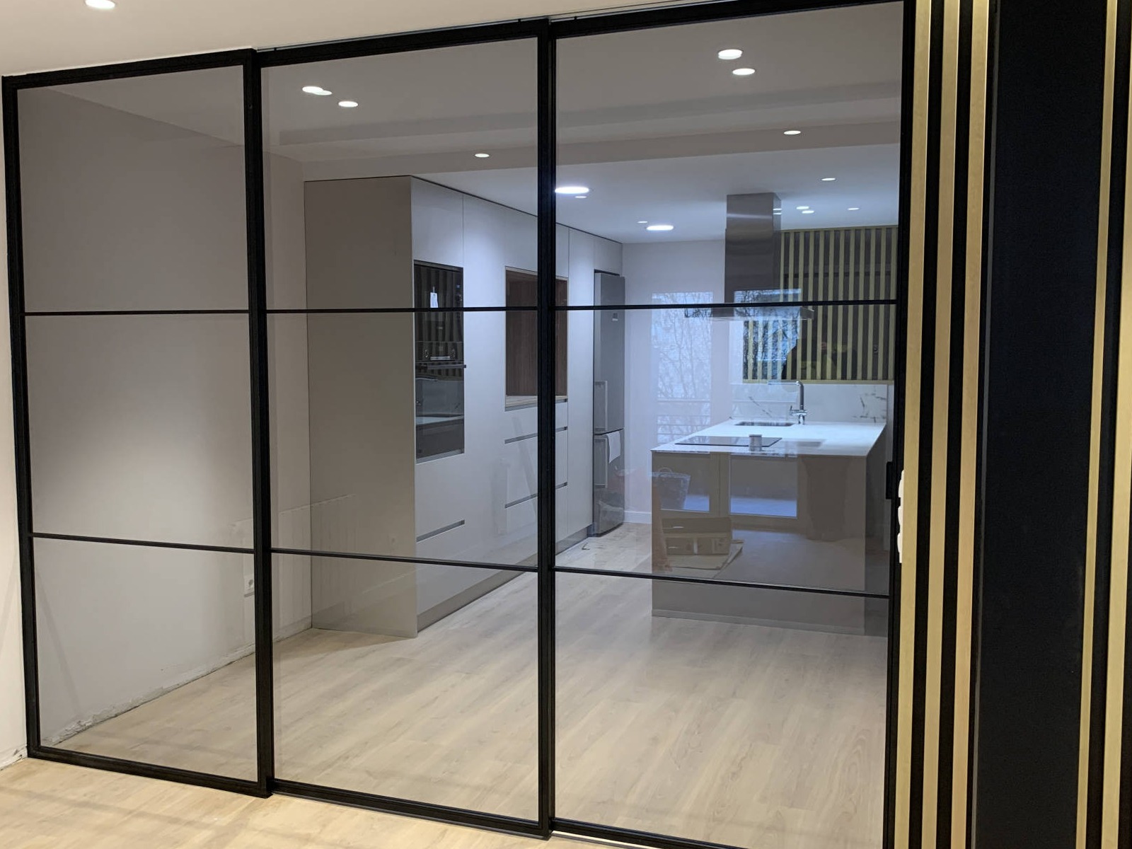 Espacio interior moderno con un sofá gris y cojines decorativos, acompañado de una mesa de centro de madera de diseño minimalista. Al fondo, un cerramiento de metal negro con cristal separa la sala de un área de trabajo o lectura. La decoración incluye estanterías de madera flotantes en la pared blanca, adornadas con pequeños objetos y marcos. El ambiente es acogedor, con un diseño contemporáneo y elementos naturales como una planta en maceta y cortinas blancas que aportan luminosidad.