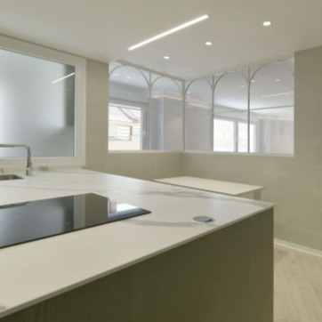 Vista lateral de una cocina moderna con encimera de mármol blanco y una placa de inducción negra. Al fondo, una puerta corredera de cristal opaco junto a ventanas decorativas acristaladas con diseño de arcos, iluminadas por luces empotradas en el techo, en un espacio amplio y minimalista.