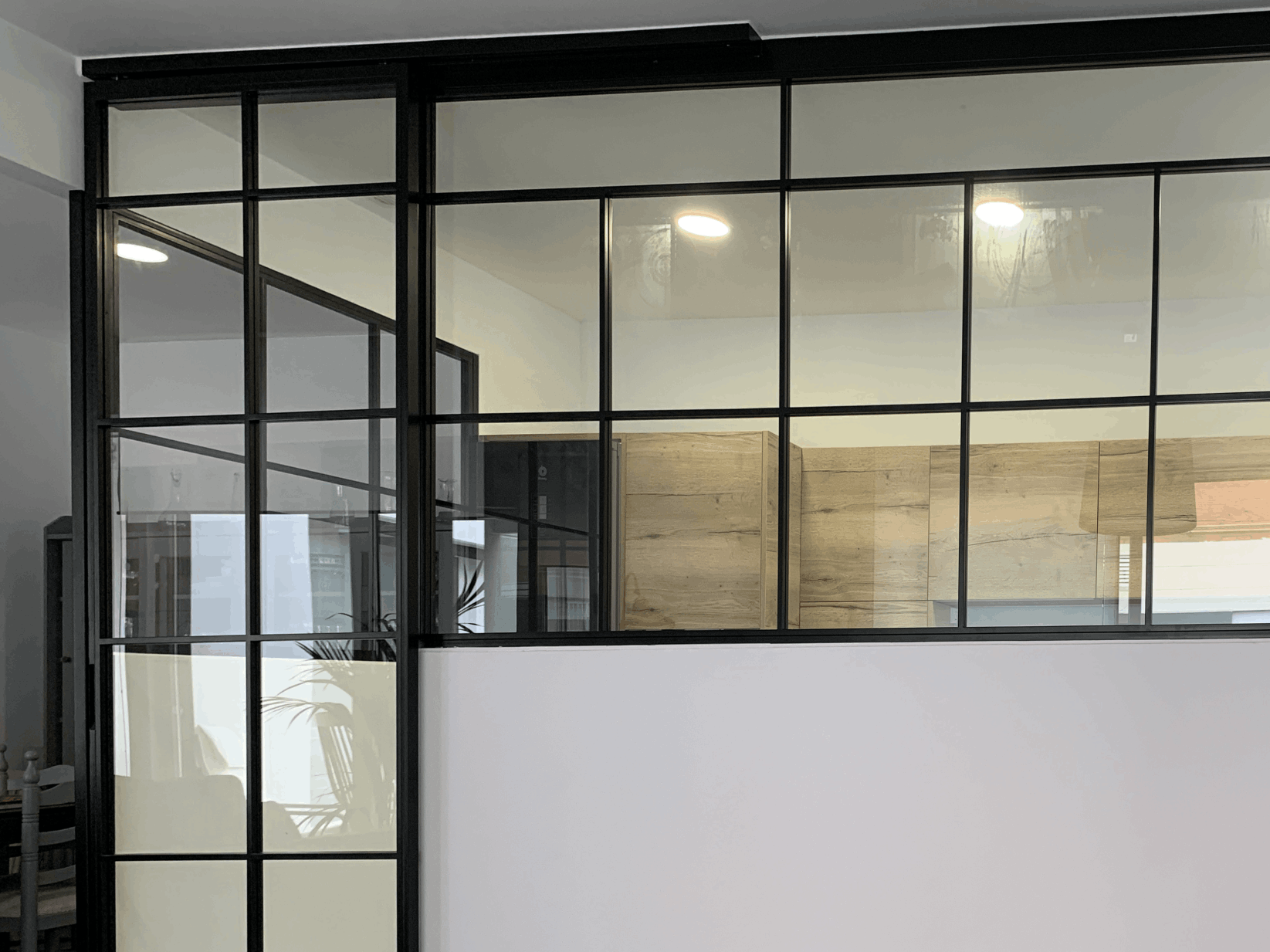 Espacio interior con cerramientos de metal y cristal que separan una sala amplia de una cocina moderna. El suelo es de madera clara, con paredes en tonos neutros y una iluminación empotrada en el techo. Destacan paneles decorativos negros con líneas doradas en el lateral derecho, aportando un diseño elegante y contemporáneo.