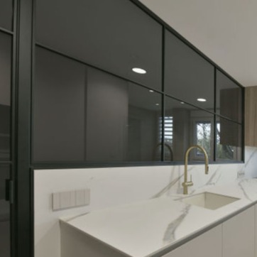 Detalle de una cocina moderna con encimera de mármol blanco con vetas grises, equipada con un fregadero integrado y un grifo dorado de diseño elegante. Al fondo, un cerramiento metálico negro con cristales que separa la cocina de otra estancia, destacando líneas limpias y un estilo contemporáneo.