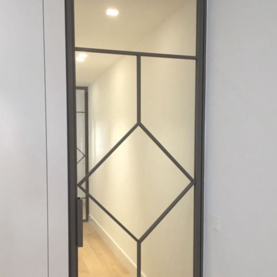Puerta corredera de diseño moderno con estructura metálica negra y cristal, destacando un diseño geométrico con formas de rombo. La puerta separa un pasillo iluminado con suelo de madera clara, aportando un estilo contemporáneo y elegante al espacio.