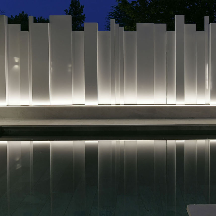 Vista nocturna de una piscina con un diseño moderno y minimalista. Al fondo, un cerramiento compuesto por paneles verticales iluminados con luz indirecta crea un efecto visual elegante y sofisticado. Los paneles, en tonos claros, reflejan su luz en el agua de la piscina, añadiendo profundidad y un ambiente tranquilo. En el exterior, se aprecian árboles y un cielo oscuro que enmarcan el entorno, reforzando la atmósfera relajante del espacio.