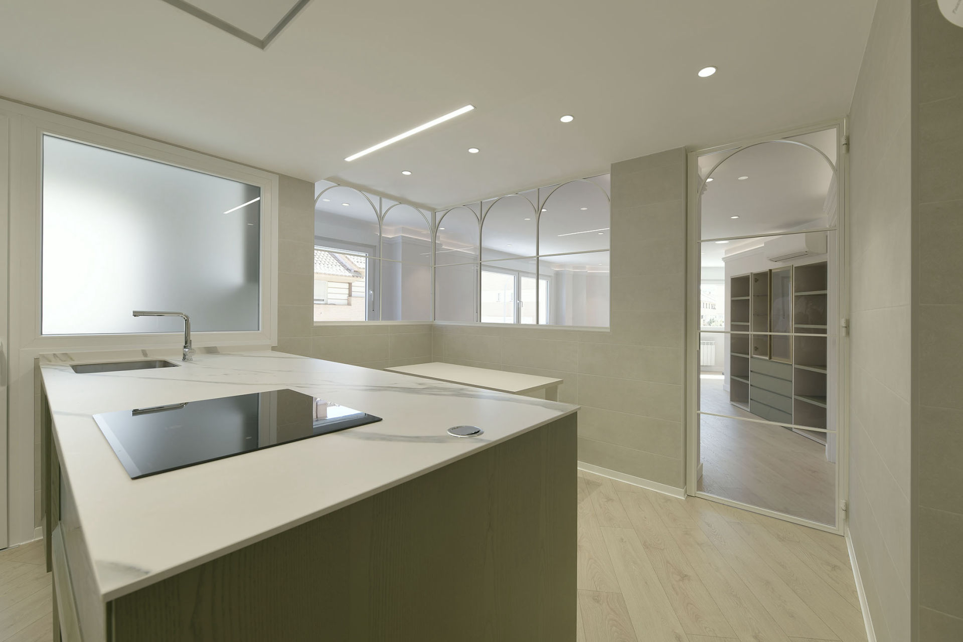 Cocina moderna con diseño minimalista y acabados de alta calidad. Cuenta con una encimera de mármol con fregadero integrado y una placa de inducción. Las paredes son de tonos neutros y destacan los cerramientos interiores de cristal con marcos metálicos curvados, que separan el espacio de otras estancias manteniendo una sensación de amplitud y luminosidad. El suelo es de madera clara, aportando calidez al ambiente, y se aprecian detalles arquitectónicos elegantes como arcos en las ventanas y puertas.
