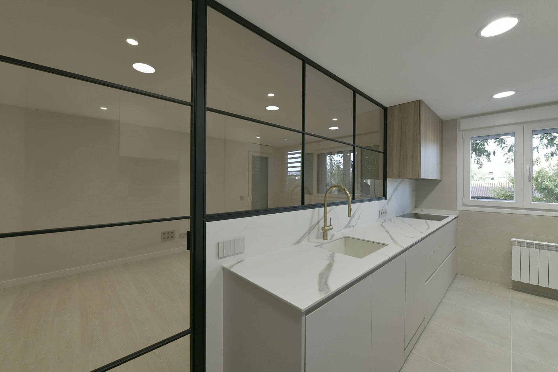 Cocina moderna con encimera de mármol blanco de vetas grises y fregadero integrado. Destaca un grifo dorado de diseño elegante que añade un toque sofisticado al espacio. Los muebles son de líneas rectas y minimalistas en color blanco, con un armario superior en acabado de madera clara. La cocina está separada de otra estancia por un cerramiento de cristal con marcos metálicos negros, que aporta amplitud y luminosidad al ambiente. En el fondo, una ventana amplia permite la entrada de luz natural, complementando el diseño limpio y funcional del espacio.