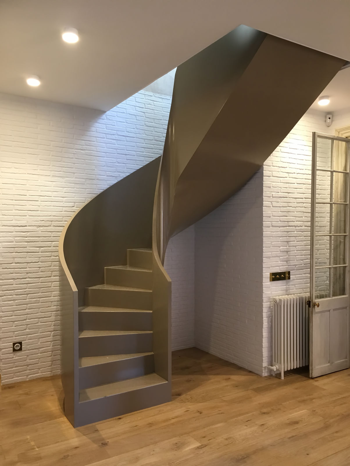 Escalera metálica de diseño moderno y minimalista, con un acabado curvo y liso en color dorado mate. La escalera se encuentra instalada en el interior de una vivienda con paredes de ladrillo blanco, suelo de madera cálida y una puerta clásica de madera con cristal. La estructura destaca por su elegancia y funcionalidad en un espacio bien iluminado.