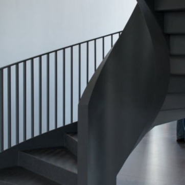 Escalera de caracol moderna hecha de metal oscuro con diseño minimalista. Los escalones y la barandilla presentan líneas limpias y rectas, resaltando un estilo contemporáneo. Fondo blanco liso que destaca la estructura.
