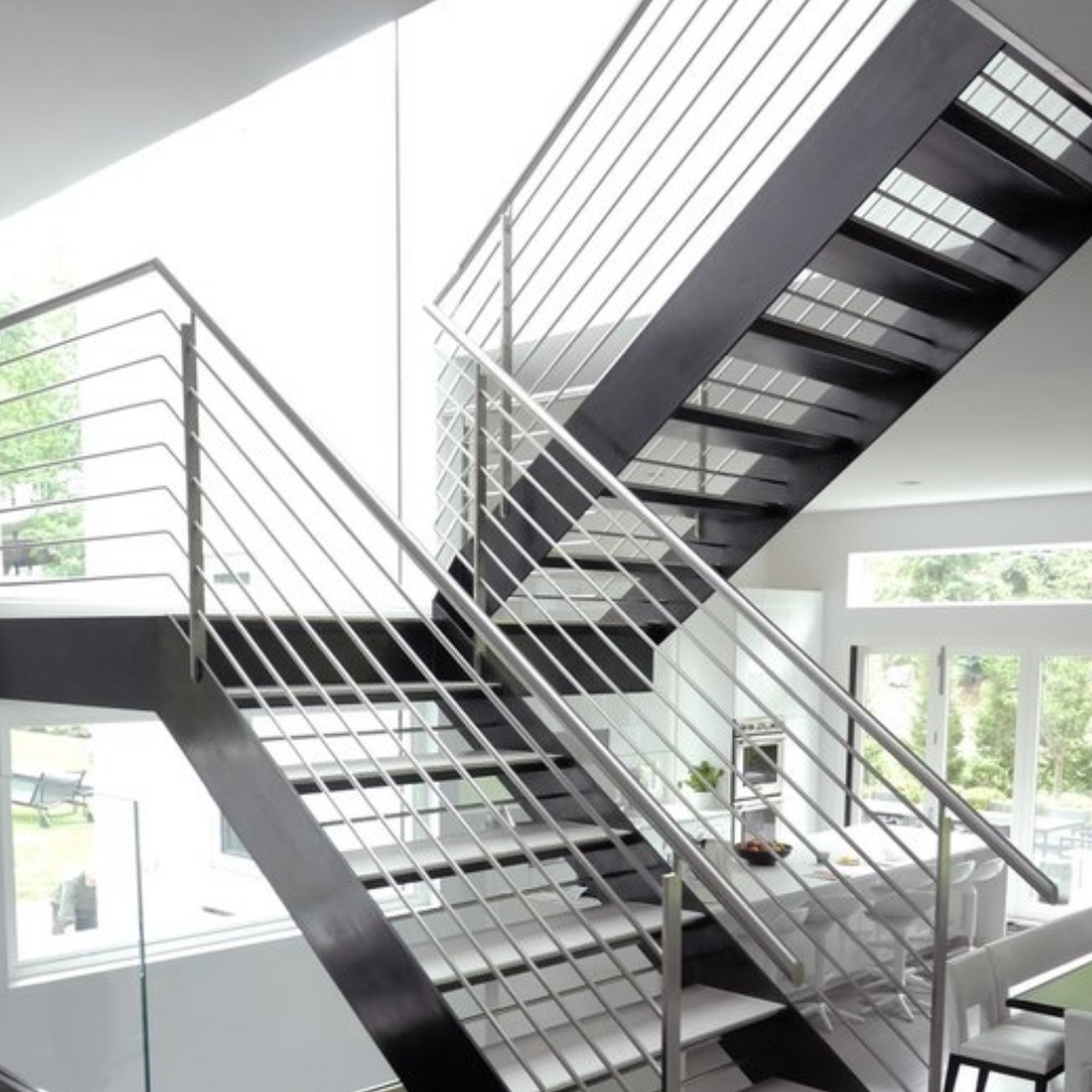 Interior de una vivienda con una escalera de diseño minimalista hecha de metal oscuro y escalones abiertos. La pared está pintada en tonos grises, con un techo de estilo industrial que muestra el acabado de hormigón. Una puerta blanca destaca en el fondo, junto a un suelo de madera clara que aporta contraste al espacio.