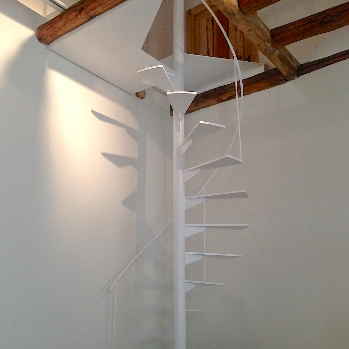 Escalera moderna de estructura metálica con escalones rectos y barandilla de cables horizontales en acero inoxidable. El diseño minimalista complementa un espacio interior luminoso, con paredes blancas y grandes ventanales que permiten la entrada de luz natural. Al fondo se observa mobiliario contemporáneo y detalles decorativos.