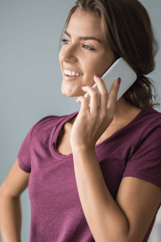 La imagen muestra a una mujer joven sonriendo mientras habla por teléfono móvil. Lleva una camiseta de color morado oscuro y su cabello está recogido de forma casual. Su expresión refleja alegría y confianza, y su postura relajada transmite una sensación de comodidad y naturalidad. El fondo es neutro, lo que centra la atención en la mujer y en su interacción con el dispositivo, ideal para representar temas de comunicación, tecnología o momentos cotidianos positivos.