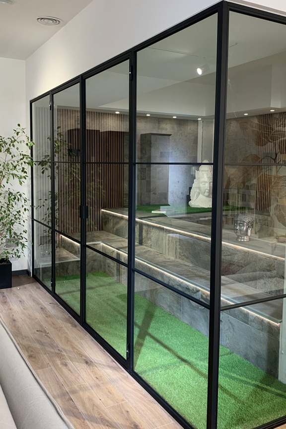 Espacio interior con diseño moderno que incluye un jardín zen con suelo de piedras negras, un árbol ornamental de hojas rojizas y grandes ventanales de vidrio que permiten vistas al exterior con vegetación. La combinación de elementos naturales y estructuras negras crea un ambiente tranquilo y elegante.